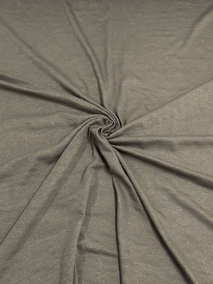 A close-up of a piece of Super Cheap Fabrics' Linen Knit - Smokey Olive - 150cm fabric with a slight sheen, arranged in a spiral pattern at the center. The folds radiate outward, creating the dynamic appearance characteristic of textured knit fabrics.