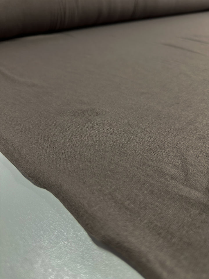 A close-up view of a roll of Super Cheap Fabrics' "Linen Knit - Smokey Olive - 150cm" unrolled on a flat surface. The material appears smooth with slight undulations.