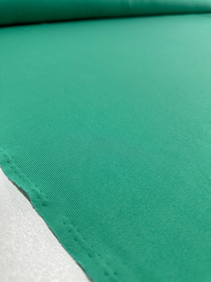 A close-up image shows a smooth, green stretch fabric rolled out on a flat surface. The Rib Knit - Peacock Green - 147cm from Super Cheap Fabrics has a slight sheen and a fine texture, with visible stitching along the bottom edge. The background is neutral, emphasizing the fabric's vibrant green color.