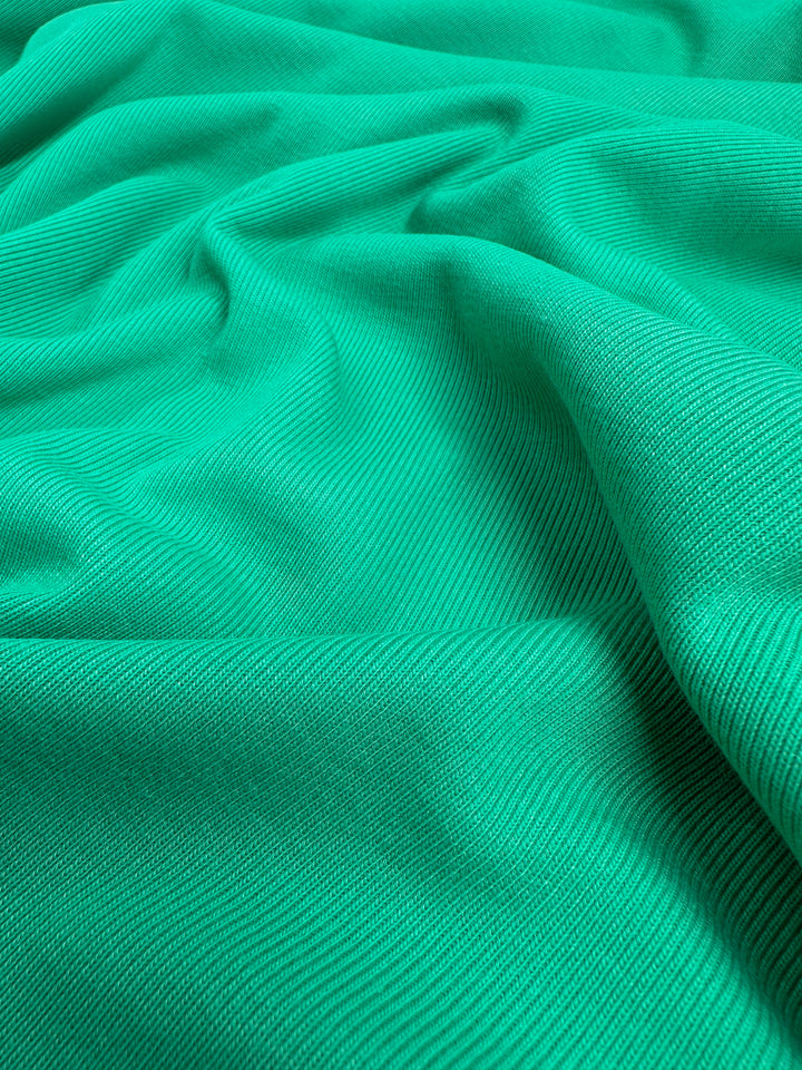A close-up photograph of a piece of vibrant green  Rib Knit - Peacock Green - 147cm from Super Cheap Fabrics. The fabric appears slightly wrinkled and draped, displaying a ribbed texture that creates subtle shadows and highlights.