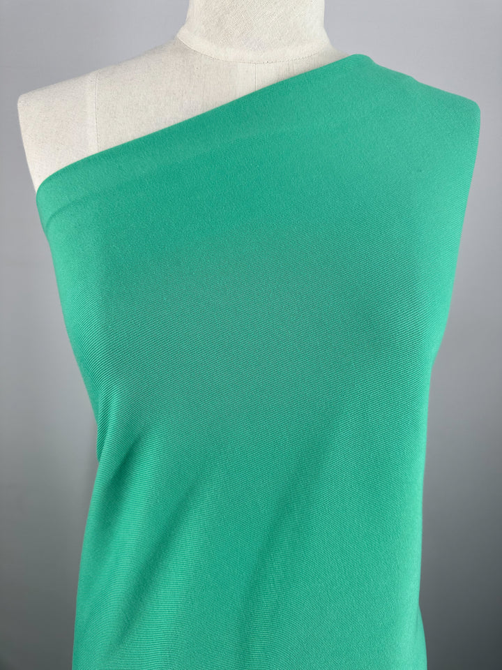 Close-up of a mannequin torso draped in a Rib Knit - Peacock Green - 147cm garment from Super Cheap Fabrics. The lightweight linen knit fabric appears to be light and stretchy, neatly fitted around the body. The background is plain and gray.