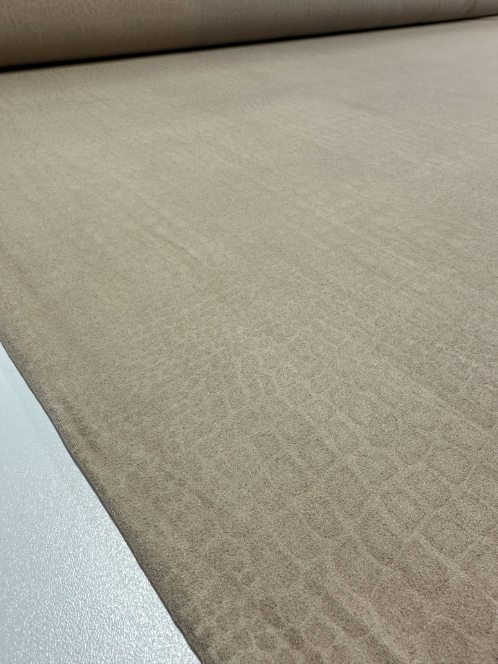 A close-up view of a large roll of light brown heavy weight fabric with a subtle textured pattern, laid out flat on a smooth, white surface. The texture resembles delicate, irregular geometric shapes, giving the Designer Faux Suede - Pepper Croc - 147cm from Super Cheap Fabrics a sophisticated appearance.