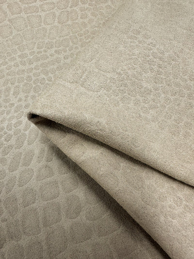 Close-up of beige textured polyester fabric with a subtle pattern resembling crocodile skin. The heavy weight fabric is arranged in overlapping layers, showing both the top surface and the underside. The texture appears soft and slightly raised, providing a tactile quality. This is Designer Faux Suede - Pepper Croc - 147cm by Super Cheap Fabrics.