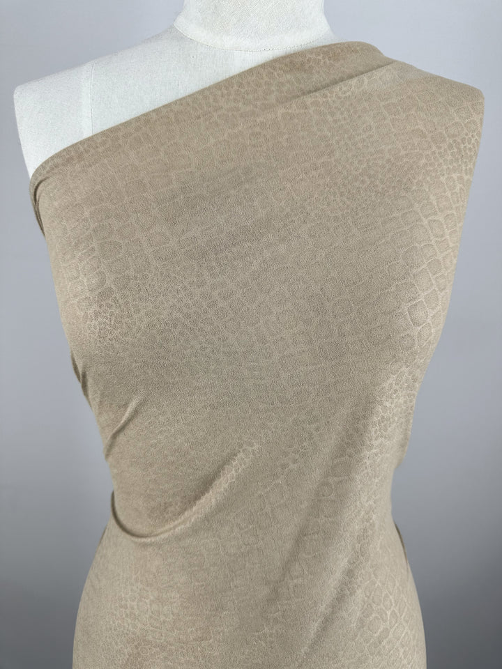 A close-up view of a beige, one-shoulder dress draped on a mannequin. The Designer Faux Suede - Pepper Croc - 147cm from Super Cheap Fabrics features a subtle alligator skin pattern, adding texture and visual interest to the garment. The background is plain and gray, drawing focus to the dress.