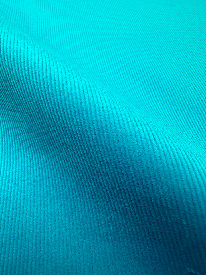 Close-up photograph of the Micro Wale Corduroy - Scuba from Super Cheap Fabrics, showcasing its blueish-green hue with parallel ribbing lines. The lighting accentuates the ridges and valleys, adding depth and detail to the fabric's surface. Ideal for winter clothing or adding a touch of texture to home décor.