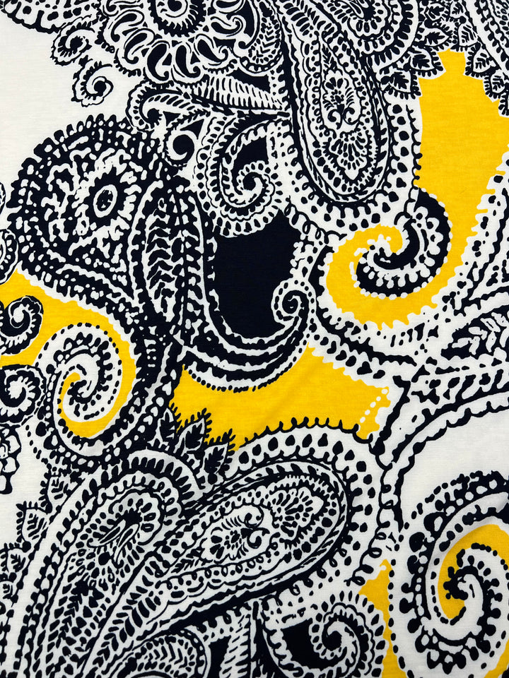 A close-up of medium weight fabric with a paisley pattern featuring intricate black and white designs on a yellow background. The Super Cheap Fabrics Printed Lycra - Mandala - 150cm material showcases detailed paisley shapes with ornate swirls and dots, creating a visually striking and complex design.