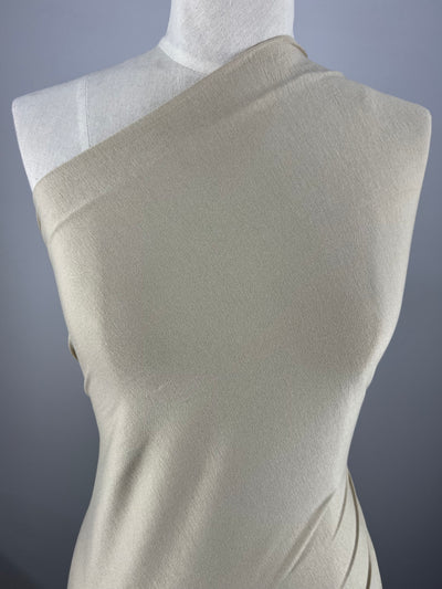 A white mannequin dressed in a cream-colored, one-shoulder garment made of medium weight fabric, Rayon Lycra - Mojave - 150cm by Super Cheap Fabrics, is positioned against a plain gray background.