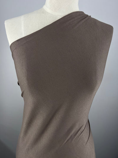 A close-up view of a mannequin wearing a brown, one-shoulder garment made from Super Cheap Fabrics' Rayon Lycra - Morel - 150cm. The smooth, fitted design showcases the elegant look of the single shoulder. The background is a plain, light grey color.