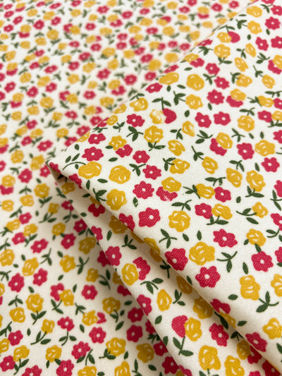 A close-up of Super Cheap Fabrics' Printed Pinwale Corduroy - Poppy - 145cm with a floral pattern featuring small, colorful flowers. The flowers are mainly pink and yellow with green leaves and stems, scattered across a white background. There are folds in the fabric, adding texture and depth to the image.