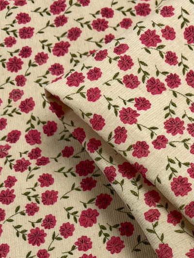 A close-up of Super Cheap Fabrics' Printed Micro Corduroy - Autumn Garden - 150cm with a beige background and a pattern featuring small pink flowers with green stems and leaves. The fabric is slightly folded, showing the continuity of the floral design, making it an exquisite choice for home decor fabric.