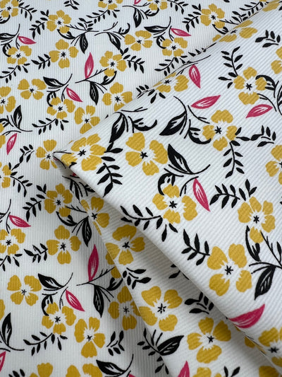 Folded Super Cheap Fabrics Printed Micro Corduroy - Sprung - 145cm with a floral pattern featuring yellow flowers, black leaves, and pink petals on a white background. The fabric appears textured with fine horizontal ribbing, perfect for autumn garden design in home decor.
