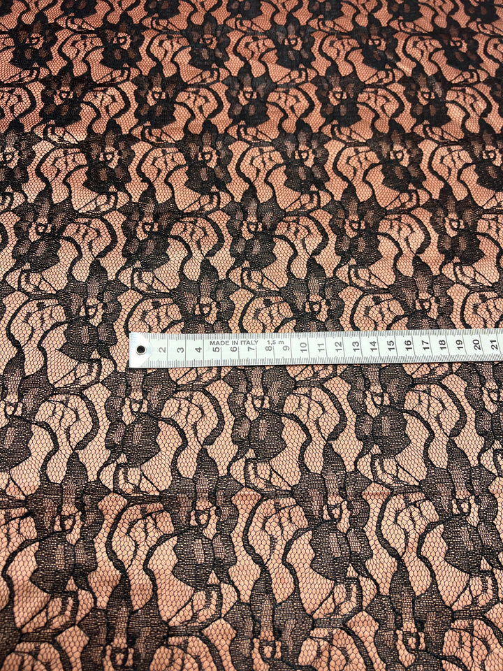 A close-up image of Super Cheap Fabrics' Lace Overlay Sateen - Copper - 150cm material laid over a nude background. A white measuring tape, marked in centimeters, is placed across the fabric horizontally, indicating the scale. The lace features an intricate repeating floral pattern.