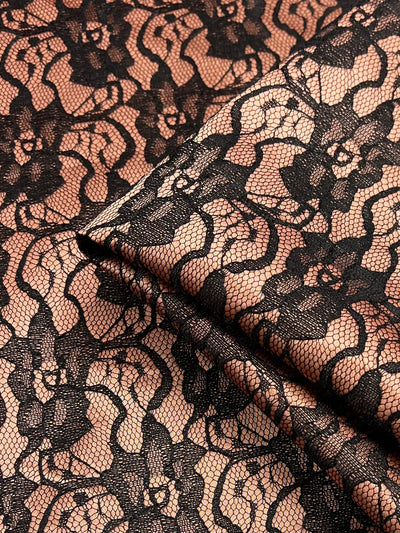 Close-up of a piece of Super Cheap Fabrics' Lace Overlay Sateen - Copper - 150cm featuring an intricate black lace pattern over a beige background. The lightweight fabric is slightly folded, showcasing the delicate floral design and the fine, woven details of the lace.