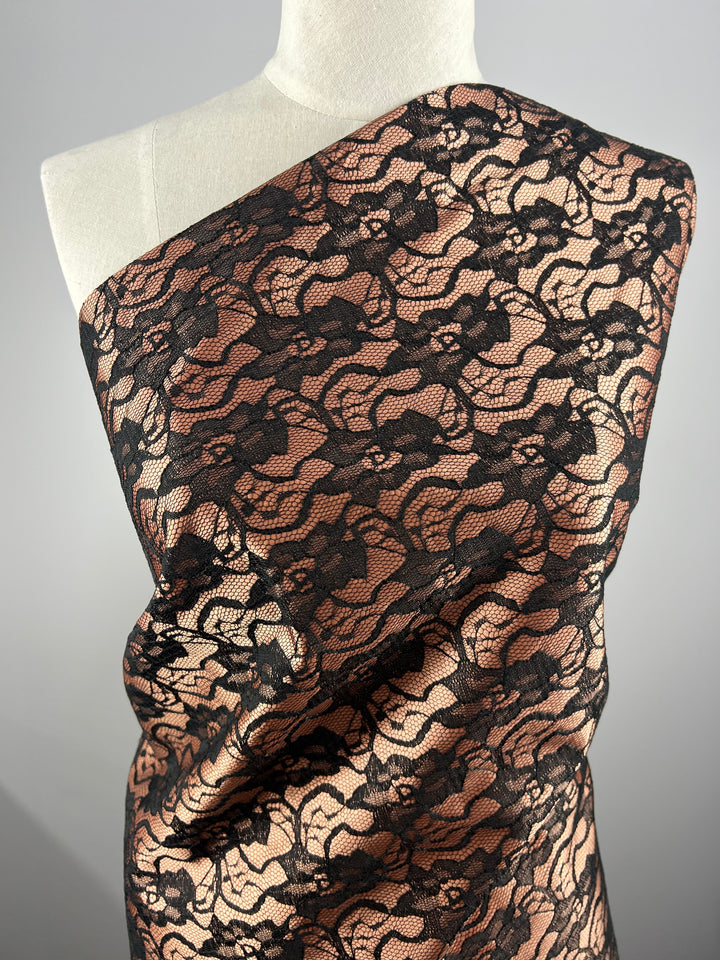 A lightweight fabric draped over a mannequin features a black lace pattern with an intricate floral design overlaying a peach background. The Lace Overlay Sateen - Copper - 150cm from Super Cheap Fabrics extends diagonally across the mannequin from the right shoulder to the left side, set against a neutral gray backdrop.