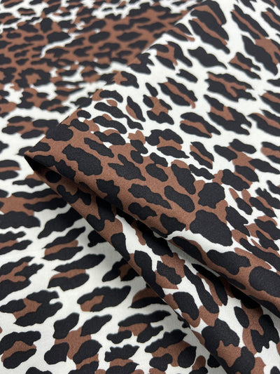 Close-up image of a lightweight cotton fabric with a leopard print pattern. The design features irregular black and brown spots on a white background. Part of the fabric is folded, showing the texture and print clearly, making it perfect for household décor. Product Name: Cotton Sateen - Striped Leopard - 148cm Brand Name: Super Cheap Fabrics