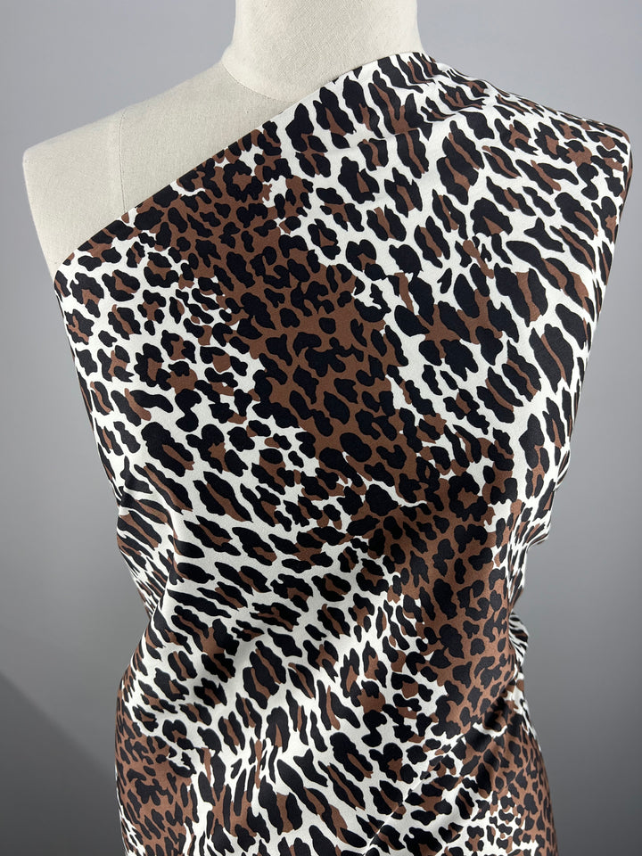 A lightweight cotton fabric with a leopard print pattern draped over a mannequin, showcasing the design. The pattern consists of large, irregular black and brown spots on a white background. The Cotton Sateen - Striped Leopard - 148cm from Super Cheap Fabrics is set against a plain gray backdrop, perfect for household décor inspiration.