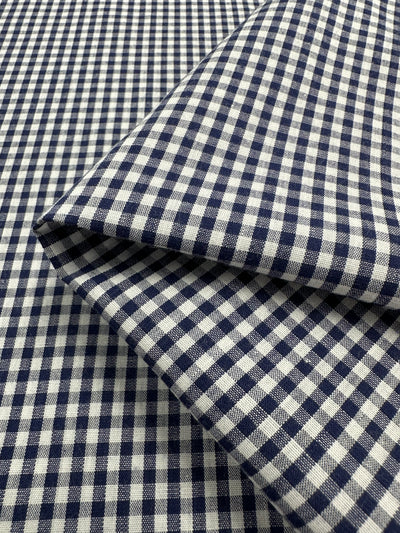 A piece of Super Cheap Fabrics' Cotton Gingham - Mini Navy - 140cm displaying a classic blue and white check pattern. The lightweight fabric is shown with a piece neatly folded over another, emphasizing the small, evenly spaced checks that create a grid-like design.