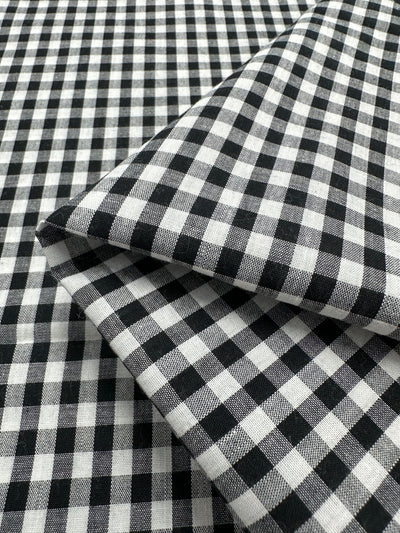 Black and white gingham fabric with a small checkered pattern is shown folded in the image. This lightweight fabric appears to be 100% cotton, ideal for creating airy and comfortable garments. The fabric is called Cotton Gingham - Mid Black - 140cm from Super Cheap Fabrics.