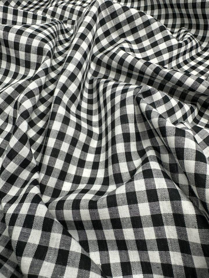 A close-up image of Cotton Gingham - Mid Black - 140cm from Super Cheap Fabrics, featuring a pattern of small squares. The 100% cotton fabric is shown in folds, creating shadows and depth, highlighting the texture and the pattern.