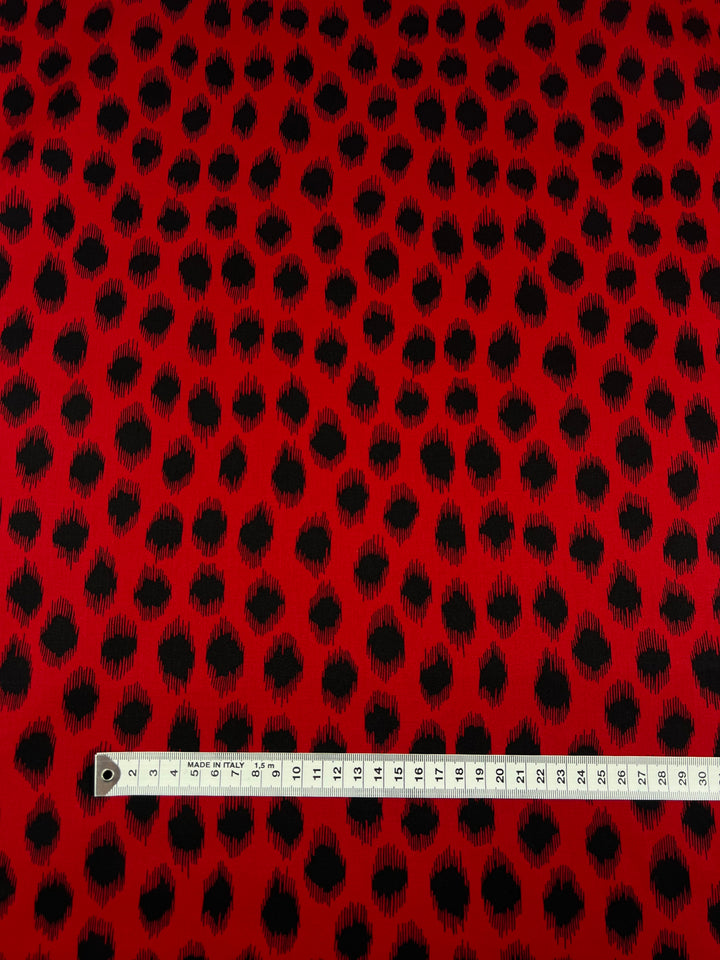 A piece of medium weight cotton fabric with a red and black irregular dot pattern is shown. A measuring tape across the bottom measures the width, marked from 0 to 50 centimeters. The product is Cotton Sateen - Red Fuzz - 150cm by Super Cheap Fabrics.