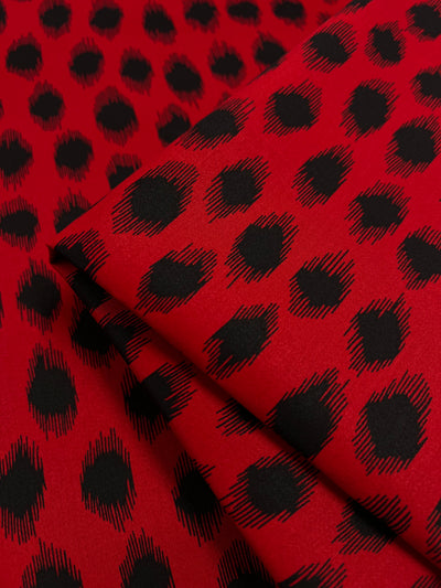 A close-up image of Super Cheap Fabrics' Cotton Sateen - Red Fuzz - 150cm with a pattern of large, irregular black dots on red. The fabric is folded over in layers, creating a layered effect with the dot pattern. The dots have a feathered texture, giving them a slightly blurred, fuzzy appearance.
