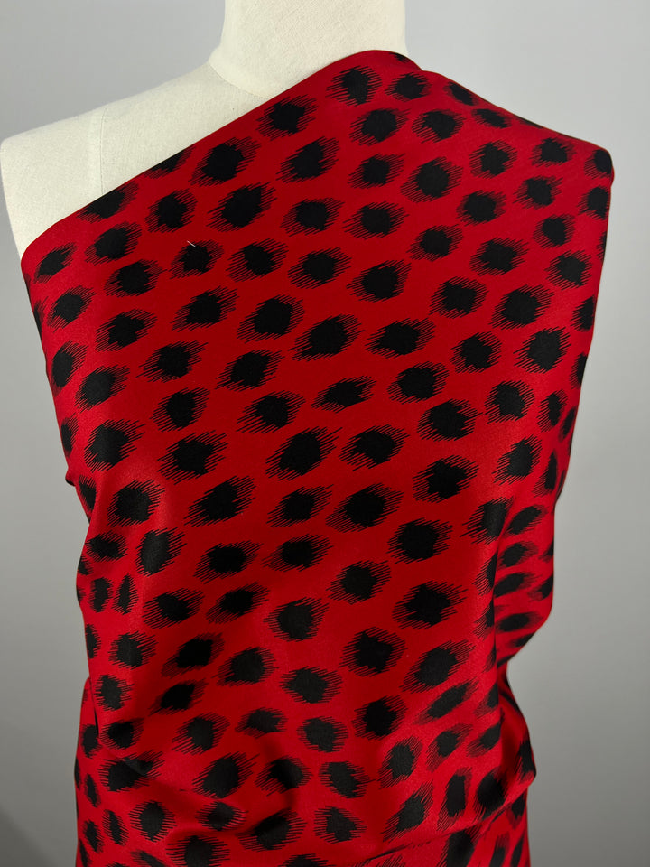 A sartorial mannequin is adorned with a vibrant red cotton fabric featuring a bold black, irregular polka dot pattern. The medium weight Cotton Sateen - Red Fuzz - 150cm from Super Cheap Fabrics is draped asymmetrically over the shoulder, highlighting its texture and design against a plain gray background.