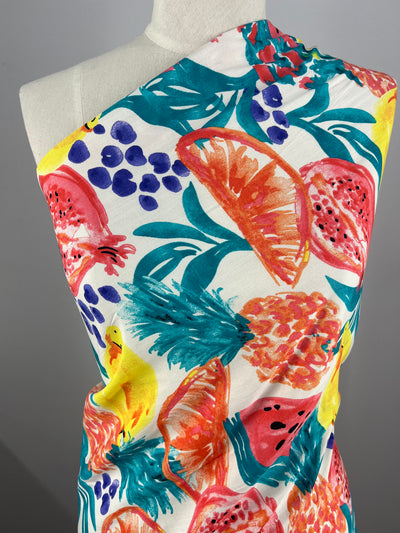 A mannequin is draped in a one-shoulder garment made from lightweight Bamboo Rayon - Fiesta - 150cm by Super Cheap Fabrics, featuring a vibrant tropical print. The design includes images of pineapples, watermelons, and various other colorful fruits and leaves on a white background.