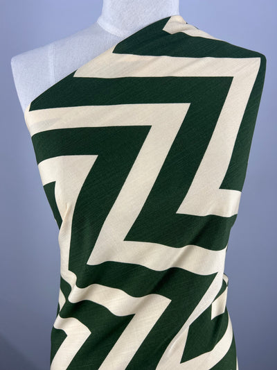 A mannequin draped with Super Cheap Fabrics' Bamboo Rayon - Zag XL - 150cm featuring a bold, green and beige zigzag pattern. The Bamboo Rayon - Zag XL - 150cm fabric is arranged diagonally across the mannequin's chest and shoulder, showcasing the geometric design. The background is a plain, light gray color.