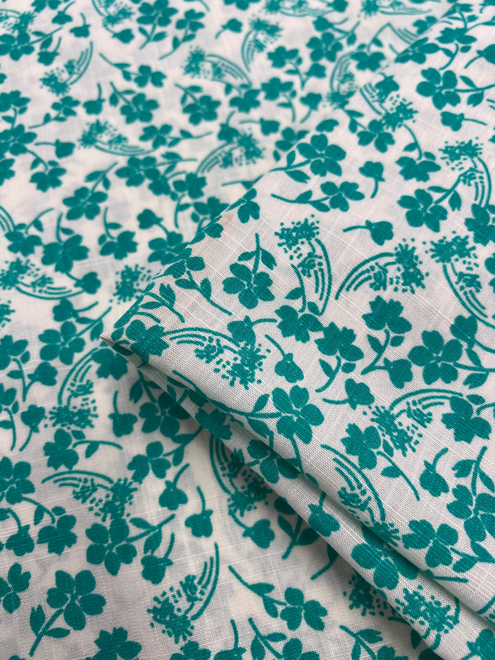 A close-up image of lightweight fabric adorned with a teal floral pattern. The design features various small flowers and leaves spread across the material, creating a delicate and intricate look. Made from Bamboo Rayon - Aqua Ditsy - 150cm by Super Cheap Fabrics, the fabric appears slightly folded in the center of the image.