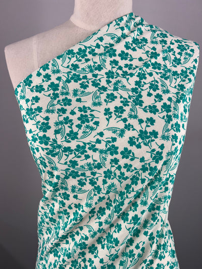 A mannequin is draped with Super Cheap Fabrics' Bamboo Rayon - Aqua Ditsy - 150cm fabric featuring a green floral pattern on a white background. The flowers and leaves are intricately designed, creating a detailed and elegant print. The Bamboo Rayon - Aqua Ditsy - 150cm fabric is arranged across the mannequin's torso diagonally.