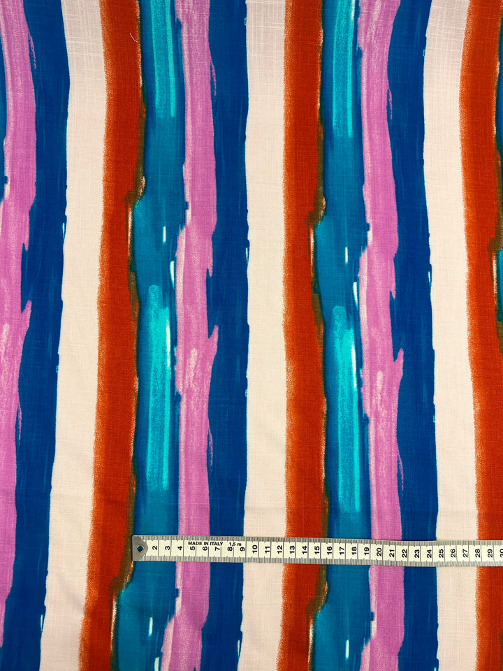 A lightweight fabric with vertical, multicolored stripes in shades of blue, teal, pink, red, and orange. A measuring tape placed horizontally across the bottom shows a length of 30 centimeters, helping to indicate the scale of the pattern. This Bamboo Rayon - Hand Stroke - 150cm material from Super Cheap Fabrics is perfect for beginner-friendly projects.