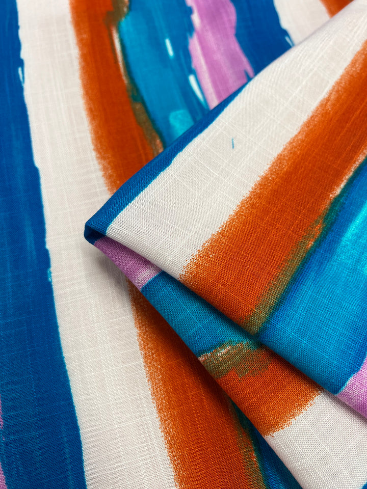 A close-up image of a lightweight, beginner-friendly fabric with a vibrant pattern. The design features bold, irregular stripes in shades of blue, orange, and purple on a white background, creating a dynamic and colorful look. The fabric is slightly folded, showcasing its texture and print. This is the Bamboo Rayon - Hand Stroke - 150cm from Super Cheap Fabrics.