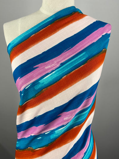 A mannequin is draped with a colorful, striped fabric featuring vibrant, horizontal bands of blue, teal, pink, orange, and white. The lightweight fabric has a watercolor brushstroke style, giving it a dynamic and artistic look. Perfect for beginner-friendly projects. This is the Bamboo Rayon - Hand Stroke - 150cm from Super Cheap Fabrics.