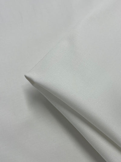 A close-up image of a neatly folded piece of Plain Sateen - Ivory - 150cm by Super Cheap Fabrics. The medium weight material appears smooth and slightly glossy, with clean lines and a minimalistic appearance. The lighting highlights the texture and sheen, making it ideal for formal wear.