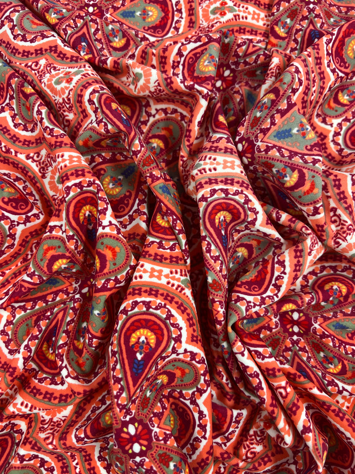A close-up view of crumpled, soft fabric with a vibrant paisley pattern in shades of red, orange, pink, and touches of green and turquoise. The intricate design includes swirls, teardrop shapes, and floral elements on the high-quality print, creating a visually rich texture perfect for multiuse projects. This is the Super Cheap Fabrics' Printed Nylon Lycra - Mirage - 150cm.