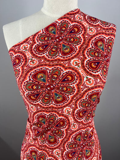 A mannequin wearing a one-shoulder garment made from Super Cheap Fabrics' Printed Nylon Lycra - Mirage - 150cm, which features a vivid red, orange, and purple floral and paisley pattern. The complex, symmetrical design includes intricate details and vibrant colors on a soft fabric background. The high-quality print creates an eye-catching appearance that is both stylish and multiuse.