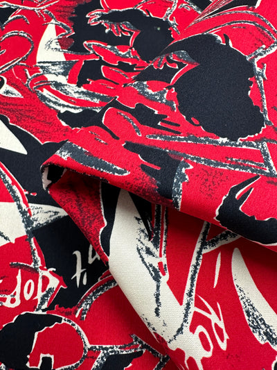 Close-up of a lightweight cotton fabric with a bold, abstract design. The pattern features large, overlapping shapes in red, black, and white, with a dynamic brushstroke-like texture. Some areas of the fabric are folded, revealing both the front and back of this versatile material ideal for household decor. This is the Cotton Sateen - Rock Roses - 150cm by Super Cheap Fabrics.