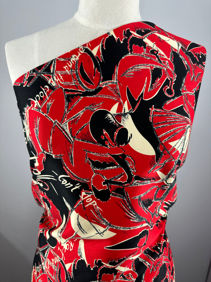 A mannequin is draped in a vibrant red, black, and white cotton fabric featuring an abstract, energetic graffiti-like pattern. The design includes bold strokes and swirls, with text and various shapes creating a dynamic visual effect. The lightweight fabric is fitted around the torso. This Cotton Sateen - Rock Roses - 150cm from Super Cheap Fabrics beautifully combines style and comfort.