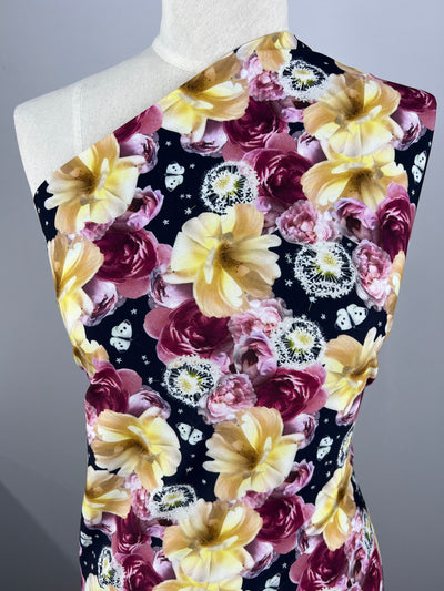 A dress form draped with Super Cheap Fabrics' Printed Lycra - Dandy - 150cm featuring a vibrant multi-colour floral pattern. The medium weight fabric design includes large, detailed flowers in shades of yellow, pink, and white against a dark background. Small white star and clock motifs are scattered throughout this Polyester Spandex blend.