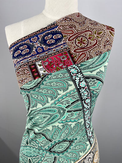A mannequin displays a single-shoulder dress featuring a vibrant, multi-colored patchwork design. The Super Cheap Fabrics Printed Lycra - Paisley Patch - 150cm includes intricate paisley, floral, and geometric patterns in shades of teal, red, brown, blue, and green, set against a light-colored background.