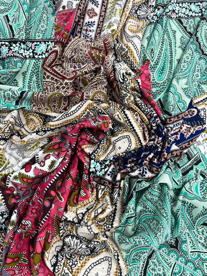A close-up image of vibrant, patterned Printed Lycra - Paisley Patch - 150cm fabrics draped and twisted together. The Super Cheap Fabrics medium weight fabrics feature intricate paisley and floral designs in various colors, including teal, brown, red, and blue. The textures and patterns blend, creating a visually rich and colorful display.