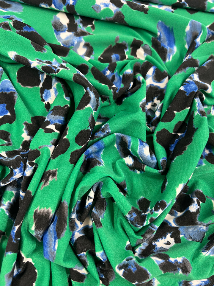 A close-up of Super Cheap Fabrics' Printed Lycra - Scattered Jungle - 150cm, a green polyester fabric with an abstract animal print pattern. The pattern features irregularly shaped black, white, and blue spots. This medium weight fabric is gathered in soft folds, creating shadows and texture, while its 2-way stretch enhances comfort and fit.