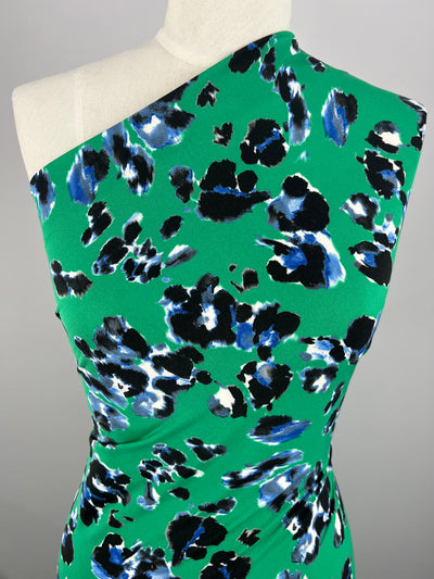 A mannequin wearing a one-shoulder dress crafted from Super Cheap Fabrics' **Printed Lycra - Scattered Jungle - 150cm**, which is made from medium weight polyester fabric with a bright green background and an abstract black, blue, and white pattern. The dress has a fitted silhouette, features a bold, artistic design, and boasts 2-way stretch for ultimate comfort. The background is plain gray.