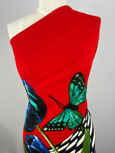 A dress form displaying a vibrant red, one-shoulder dress made from medium-heavy weight polyester fabric, adorned with a large, detailed blue and green butterfly and abstract blue flower designs. The lower part features bold black and white geometric patterns. The dress contrasts strikingly against the plain background. This striking piece showcases the Textured Knit - Red Butterfly - Panel - 85cm by Super Cheap Fabrics.