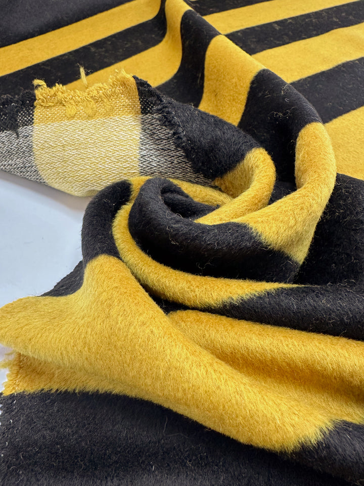 A close-up of Super Cheap Fabrics' Wool Cashmere - Honey (154cm) reveals a soft texture with bold black and yellow stripes. The fabric curls at the center, creating texture, while a slightly frayed striped end adds rustic charm and luxurious softness for year-round use.