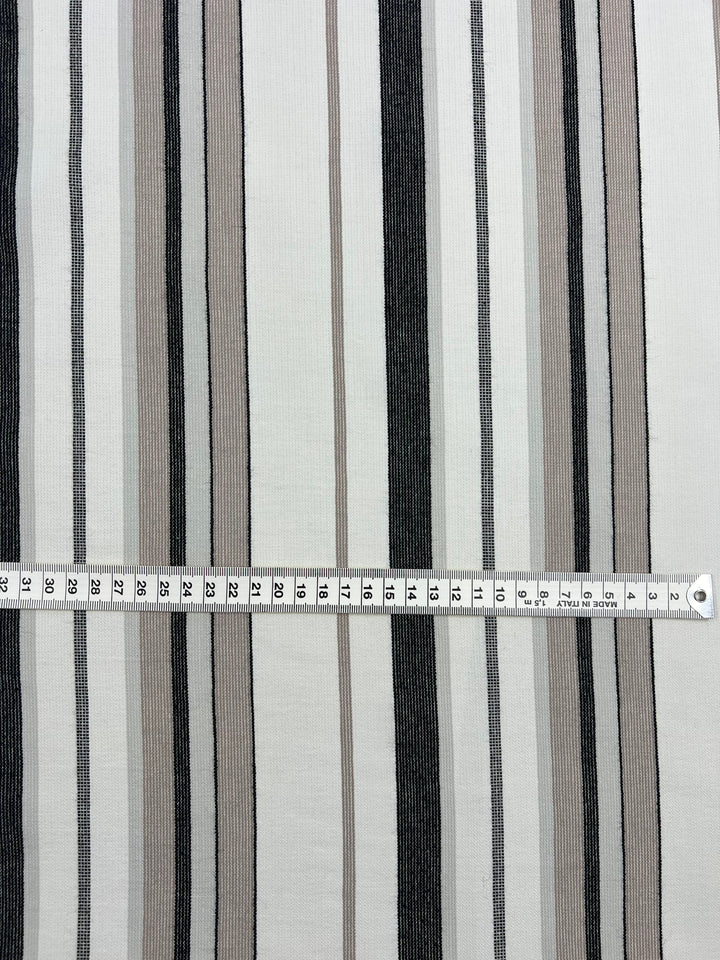 A fabric from the Super Cheap Fabrics Designer Cotton - Grey Rock Candy - 145cm with vertical black, grey, and beige stripes of varying widths, featuring a measuring tape laid horizontally across it to show measurements in centimeters. This eco-friendly fashion piece highlights our commitment to using sustainable deadstock materials.