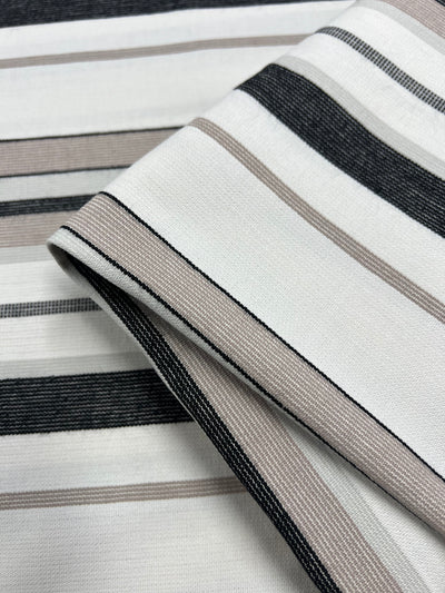 Close-up of folded Designer Cotton - Grey Rock Candy - 145cm fabric by Super Cheap Fabrics with a pattern of horizontal lines. The fabric features alternating stripes in white, black, and various shades of gray and beige. Utilizing sustainable deadstock materials, the texture appears smooth and the colors are muted, creating a subtle, elegant design for eco-friendly fashion.