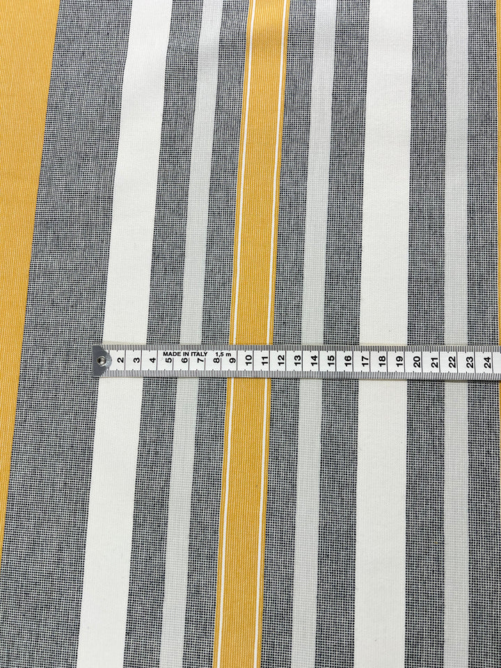 A measuring tape is placed across a fabric with vertical stripes. The eco-friendly fashion fabric features alternating black, white, and bright yellow stripes of varying widths. The measuring tape shows measurements in centimeters on the Designer Cotton - Monaco - 145cm by Super Cheap Fabrics.