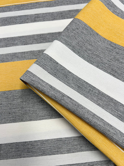 Striped fabric with a pattern that alternates between broad yellow, gray, and white horizontal stripes. The gray stripes have a fine mesh-like texture. Part of the Designer Cotton Range, this sustainable deadstock material is neatly folded on a larger piece of the same design. Introducing Designer Cotton - Monaco - 145cm by Super Cheap Fabrics.