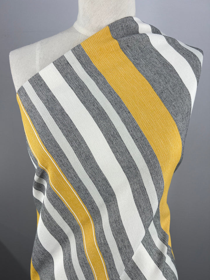A piece of fabric from Super Cheap Fabrics' Designer Cotton - Monaco - 145cm with alternating vertical stripes in yellow, white, and gray is draped over a mannequin. The mannequin is set against a plain gray background, showcasing our commitment to eco-friendly fashion made from sustainable deadstock materials.